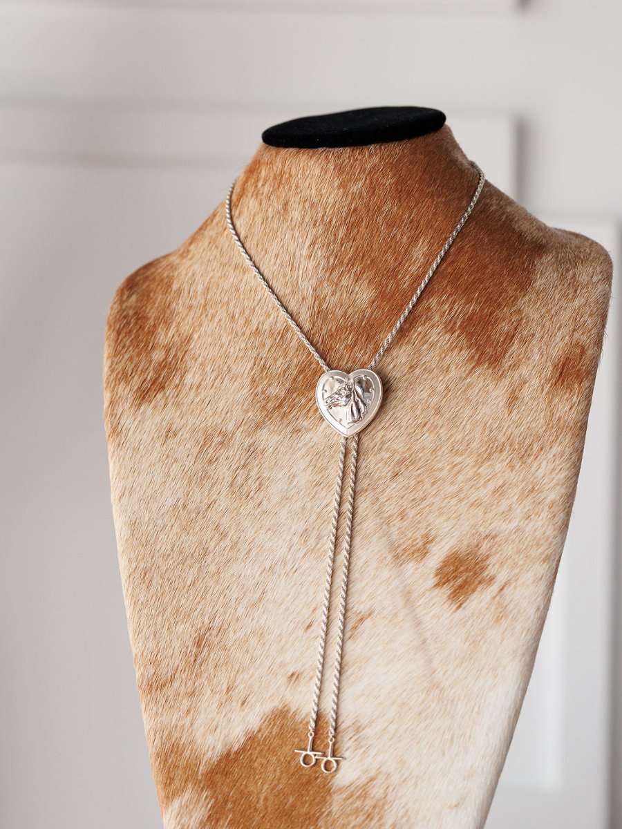 The Savannah Sevens western life{&}style Wild At Heart Bolo features a heart-shaped sterling silver pendant and dual chain strands, displayed on a cowhide-patterned stand for true Western charm.
