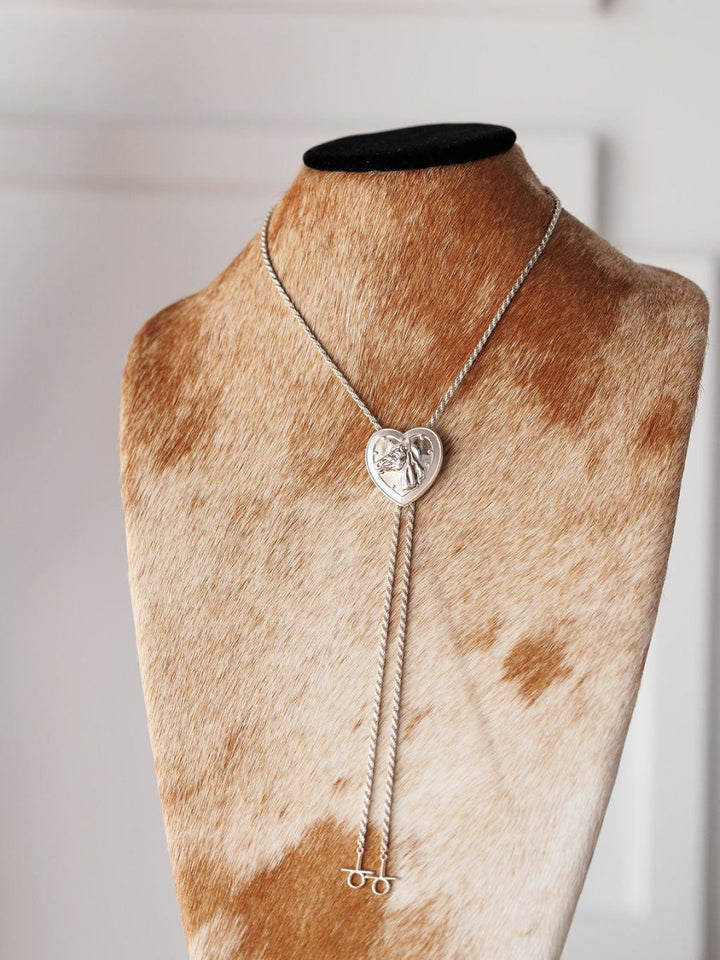 The Savannah Sevens western life{&}style Wild At Heart Bolo features a heart-shaped sterling silver pendant and dual chain strands, displayed on a cowhide-patterned stand for true Western charm.