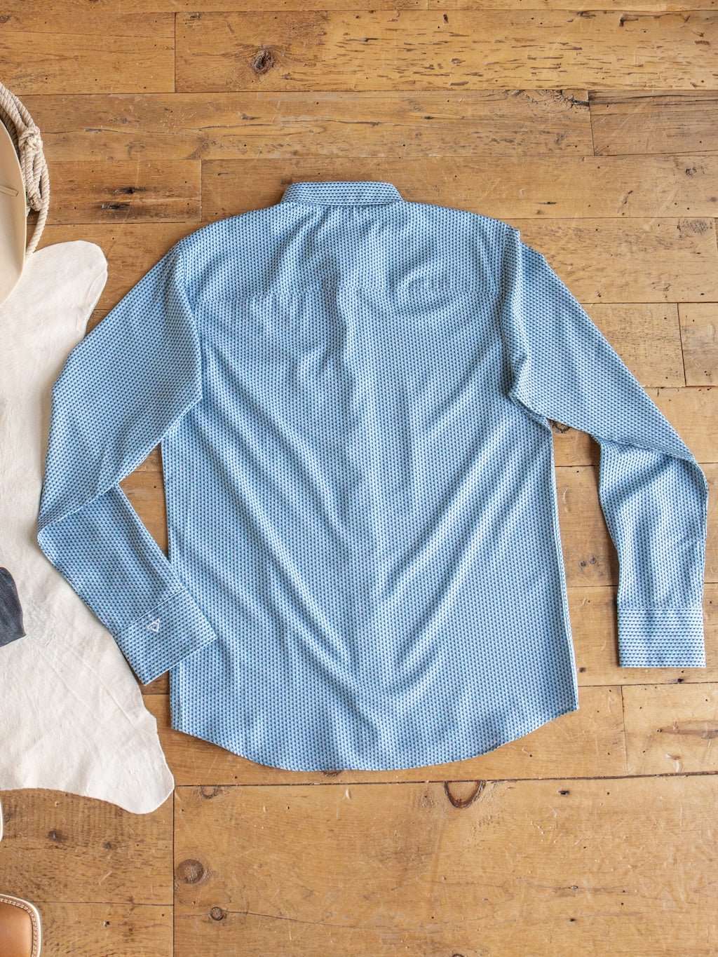 The Willsboro Wrangler Men's Blue Button Up by Savannah Sevens western life{&}style is laid flat with the back side facing up on a wooden floor, with the edges of a cowhide rug and a basket visible in the background.