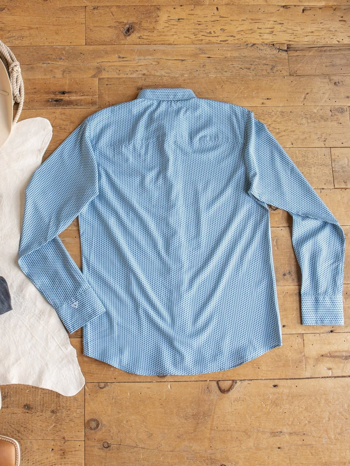 The Willsboro Wrangler Men's Blue Button Up by Savannah Sevens western life{&}style is laid flat with the back side facing up on a wooden floor, with the edges of a cowhide rug and a basket visible in the background.