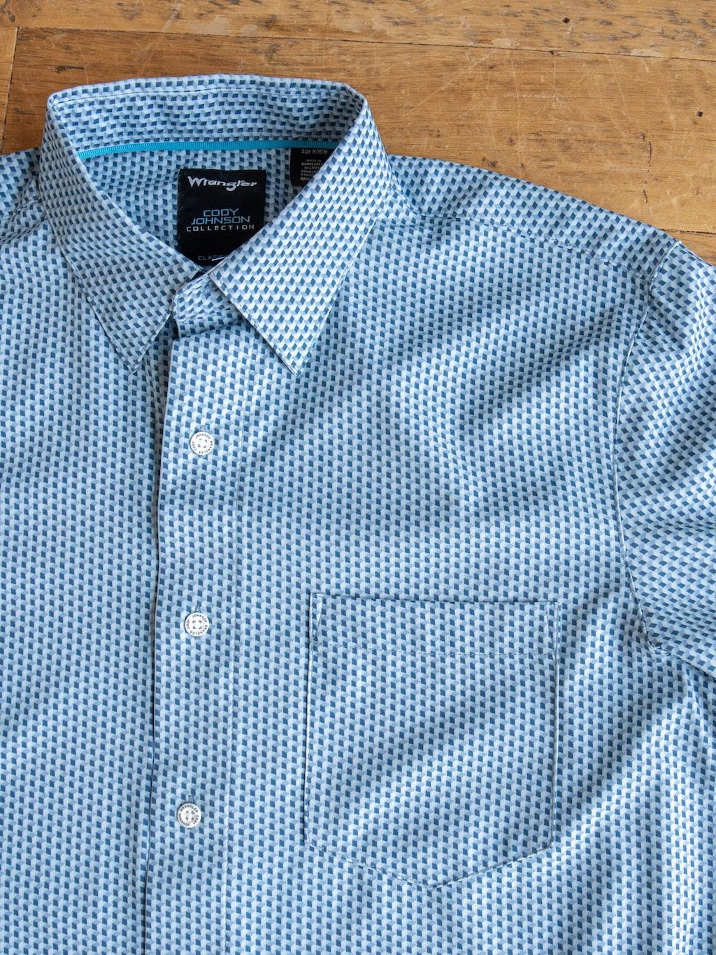 The Willsboro Wrangler Men's Blue Button Up from Savannah Sevens western life{&}style is laid flat on a wooden surface, displaying its collar, chest pocket, and buttons. This moisture-wicking shirt combines stylish geo patterns with lasting comfort.