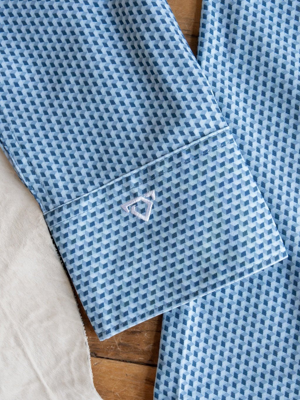 A close-up of the Willsboro Wrangler Men's Blue Button Up from Savannah Sevens western life{&}style shows its geo pattern sleeve with blue and white designs and a small embroidered geometric logo near the cuff, lying on a wooden surface next to a light-colored fabric. This moisture-wicking shirt blends style and comfort for any occasion.