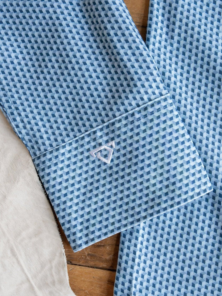 A close-up of the Willsboro Wrangler Men's Blue Button Up from Savannah Sevens western life{&}style shows its geo pattern sleeve with blue and white designs and a small embroidered geometric logo near the cuff, lying on a wooden surface next to a light-colored fabric. This moisture-wicking shirt blends style and comfort for any occasion.