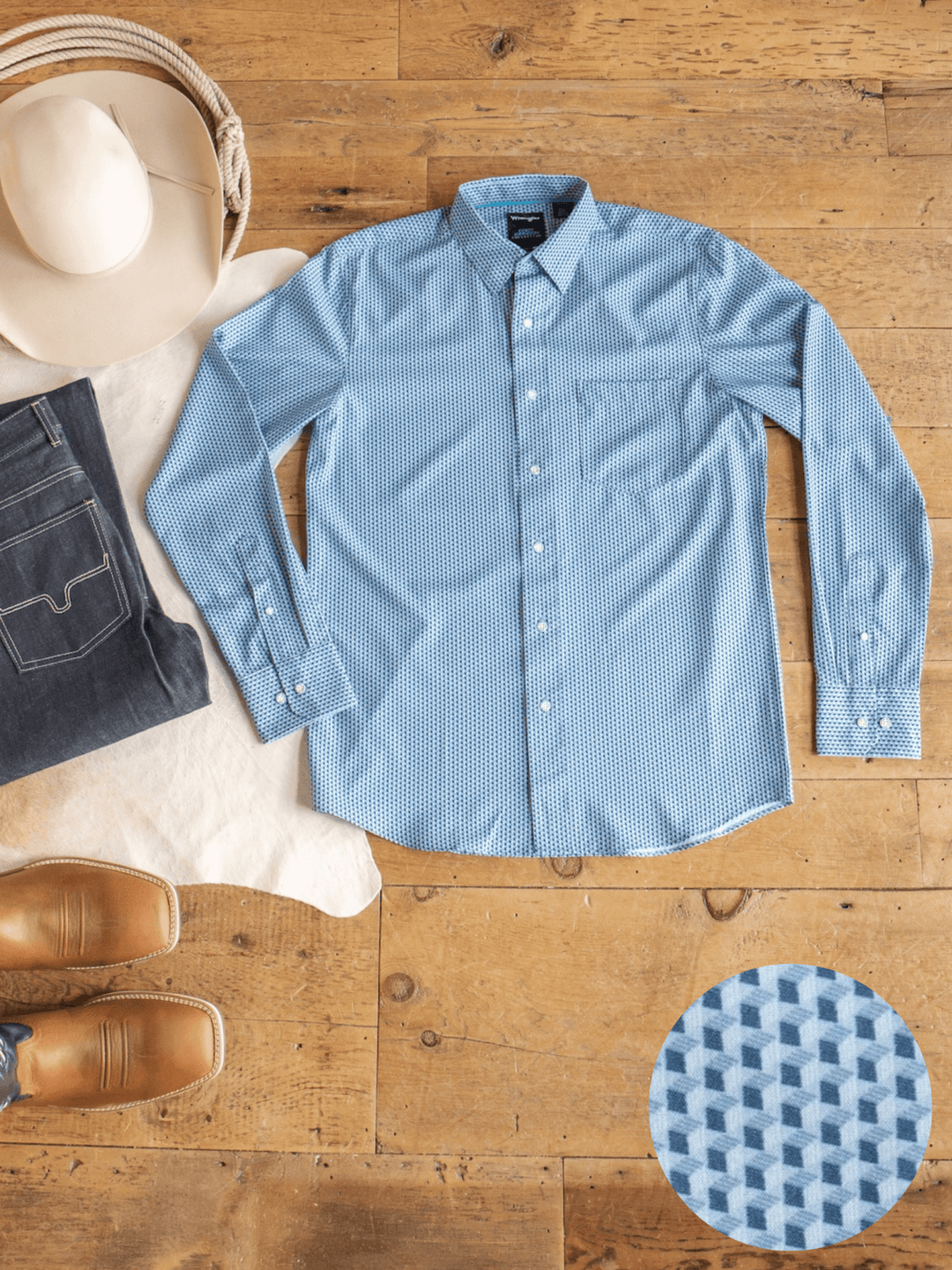 The Willsboro Wrangler Men's Blue Button Up from Savannah Sevens western life{&}style is laid flat on a wooden floor beside jeans, a cowboy hat, tan boots, and a close-up of its geometric print. This moisture-wicking button-up offers both style and comfort.