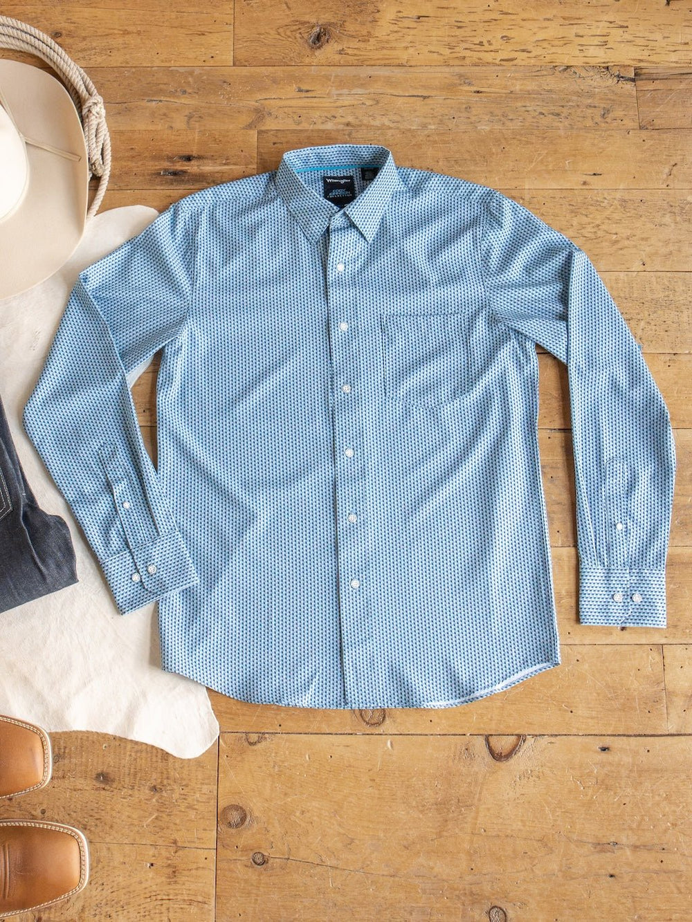 A Willsboro Wrangler Men's Blue Button Up shirt by Savannah Sevens western life{&}style, featuring a geo pattern and long sleeves, is laid flat on a wooden floor with the edge of a hat, jeans, and tan boots visible nearby.