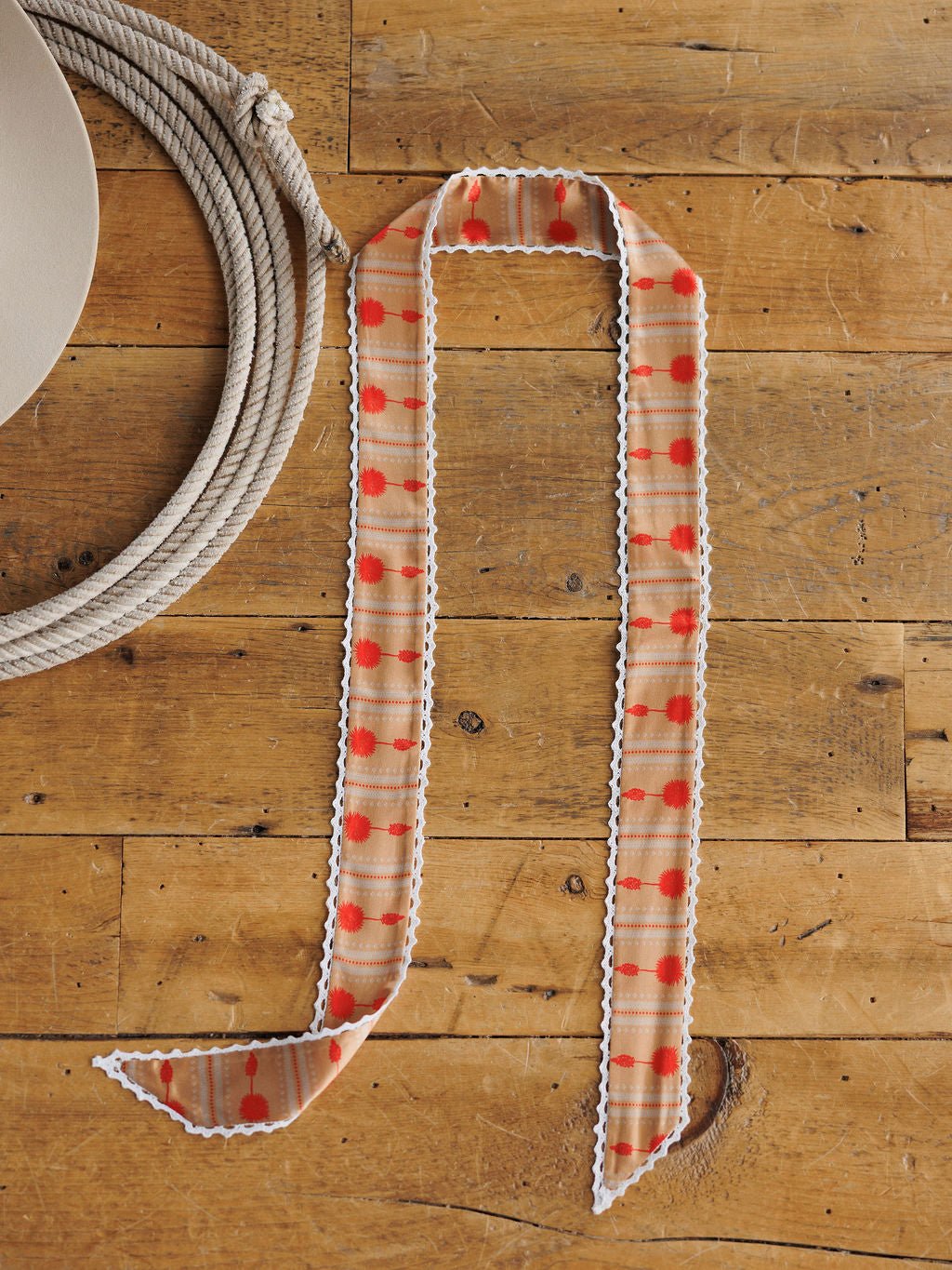 The Winnie Twilly by Savannah Sevens western life{&}style, a tan and orange checked narrow scarf with white crochet trim, is displayed on rustic wood beside a coiled rope and the brim of a beige hat.