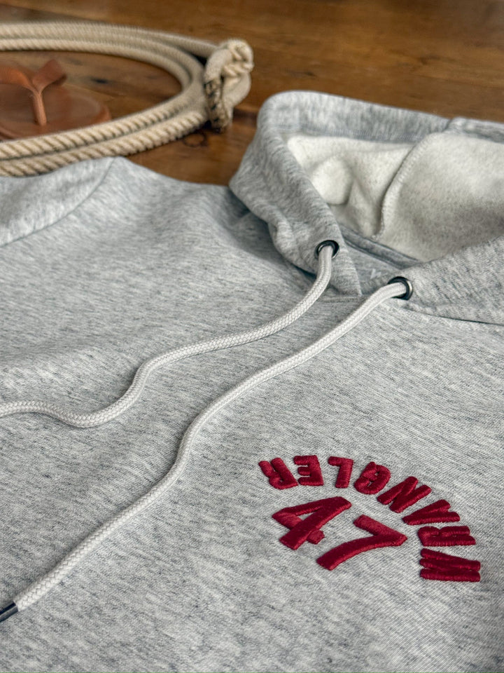 A Winnsboro Heather Grey Hoodie by Savannah Sevens western life{&}style, featuring WRANGLER 47 embroidered in red on the chest, lies on a wooden surface next to a coiled rope and a brown leather coaster.