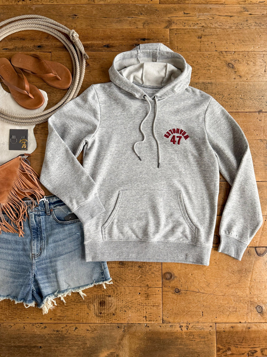 A Winnsboro Heather Grey Hoodie by Savannah Sevens western life{&}style, featuring WRANGLER 47 in red, styled with denim shorts, tan sandals, a fringed brown bag, a rope, and sunglasses laid out on a wooden floor.