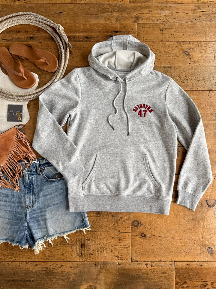 A Winnsboro Heather Grey Hoodie by Savannah Sevens western life{&}style, featuring WRANGLER 47 in red, styled with denim shorts, tan sandals, a fringed brown bag, a rope, and sunglasses laid out on a wooden floor.