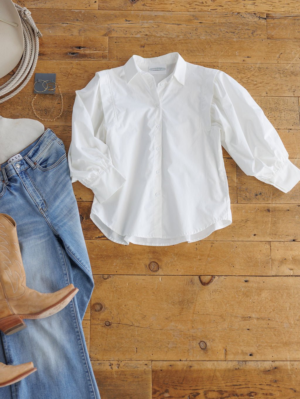 The Wiota White Cotton Button Up Top by Savannah Sevens western life{&}style is laid out on a wooden floor beside light blue jeans, tan cowboy boots, a cream hat, and silver jewelry.