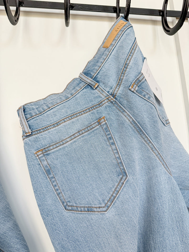The Wylie High Rise Wide Leg Jean by Savannah Sevens western life{&}style in a light wash hangs on a black metal rack, displaying the back pocket, beige waistband label, and a white size tag.