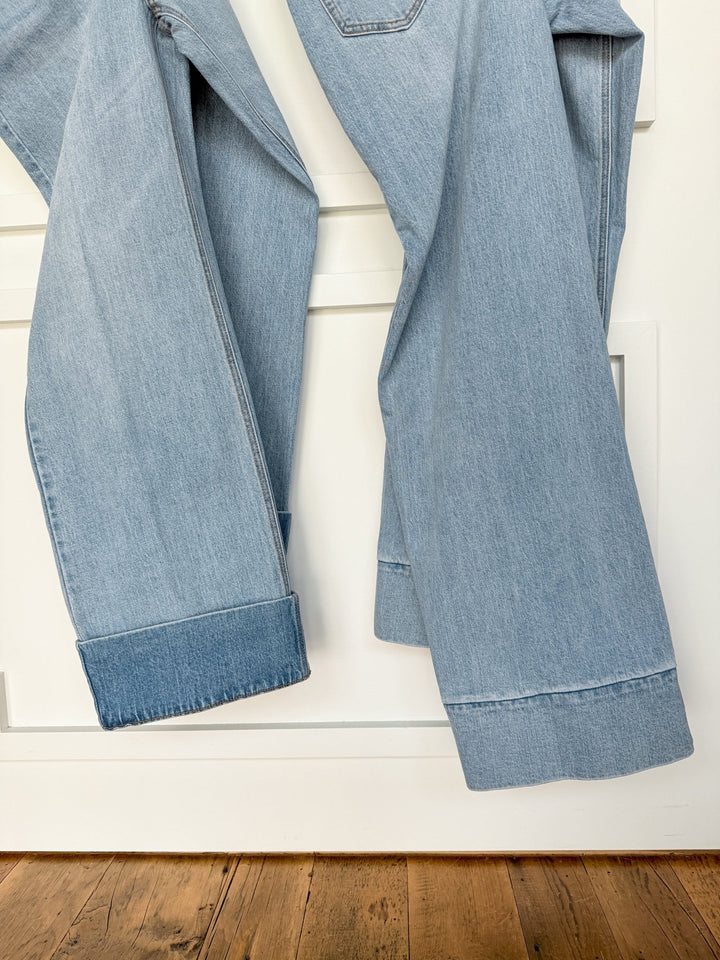 The Wylie High Rise Wide Leg Jean by Savannah Sevens western life{&}style hangs over a white door, natural light highlighting its textured light wash denim and cuffed hems for a relaxed look against the wooden floor.