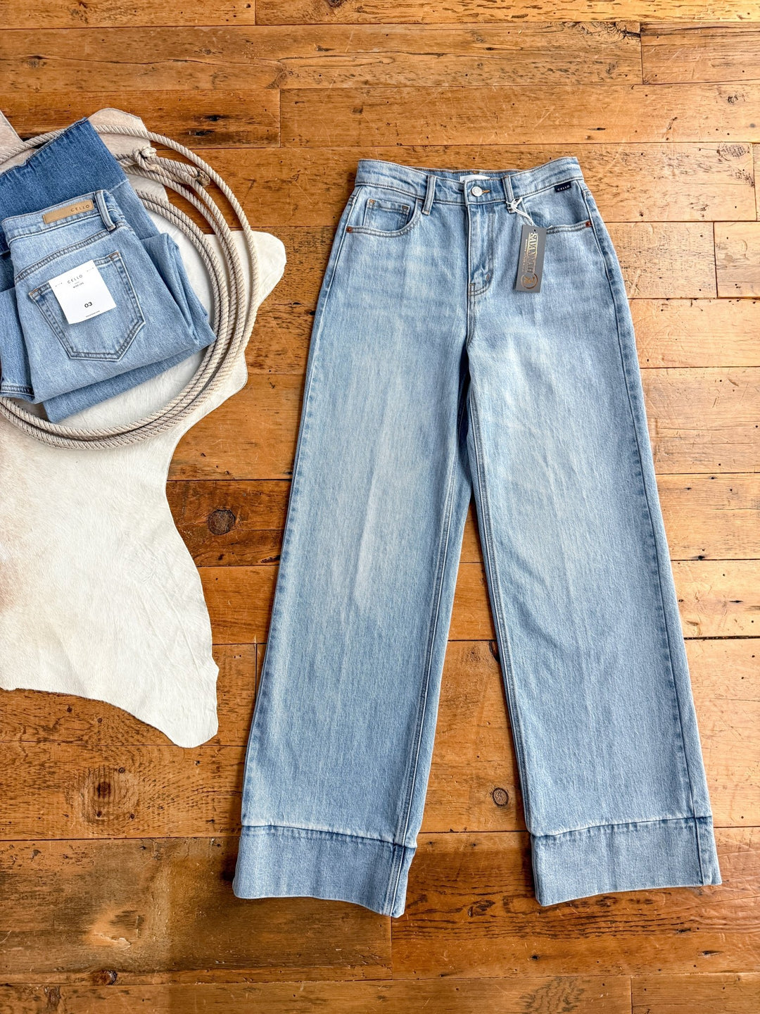 The Wylie High Rise Wide Leg Jean by Savannah Sevens western life{&}style features light wash denim and folded cuffs, laid flat on a wooden floor with tags visible. Folded jeans and a coiled rope sit nearby on a white cowhide rug.