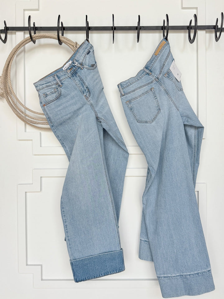 Two pairs of The Wylie High Rise Wide Leg Jeans by Savannah Sevens western life{&}style hang side by side on black hooks against a white paneled wall, with a thick rope draped on the left. Both jeans feature large cuffs at the bottom.