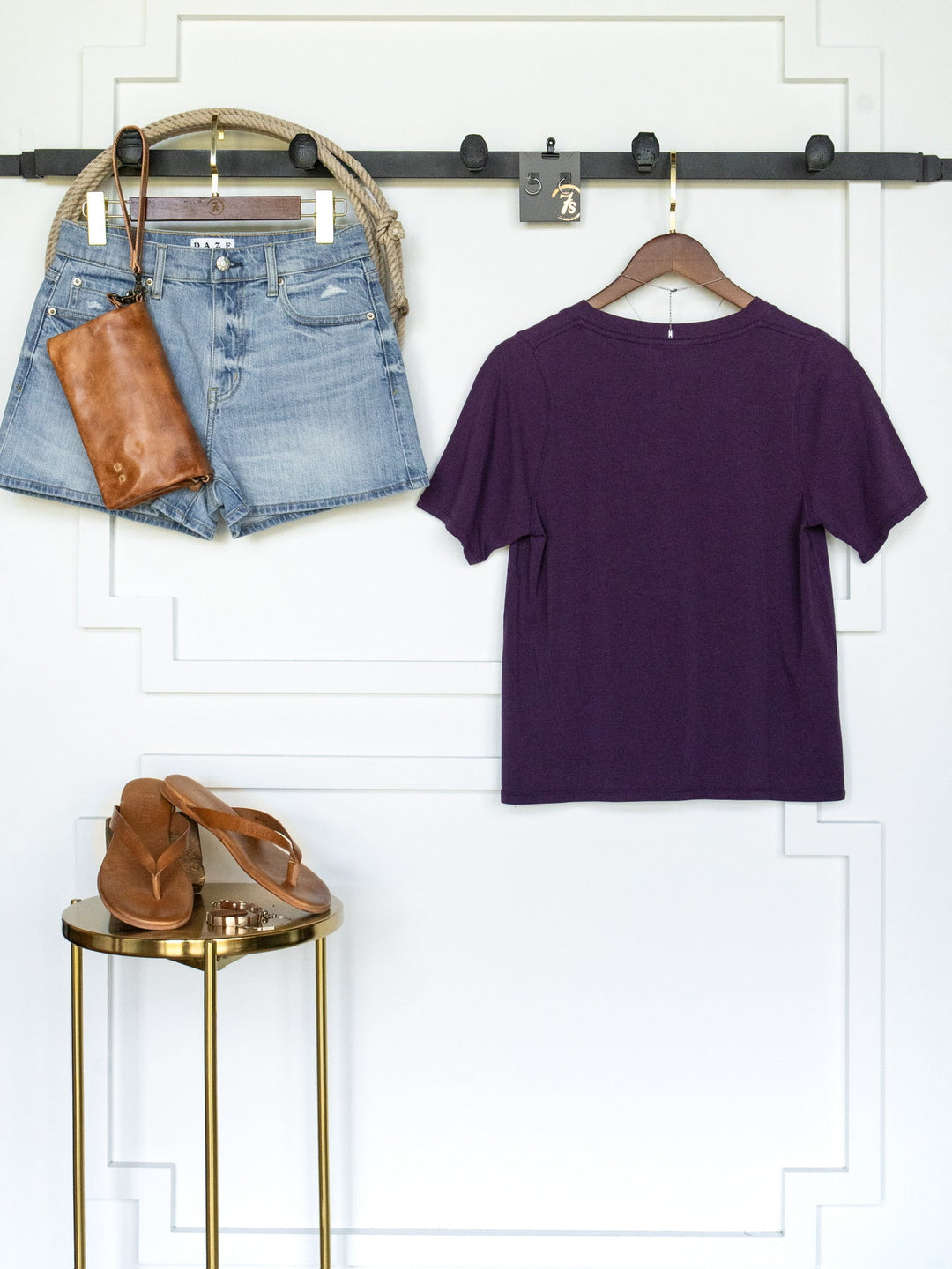 The Wymore Eggplant Basic Tee by Savannah Sevens western life{&}style hangs on a hanger, paired with denim shorts, a brown leather clutch, and sandals—all arranged neatly against a white wall with decorative trim.