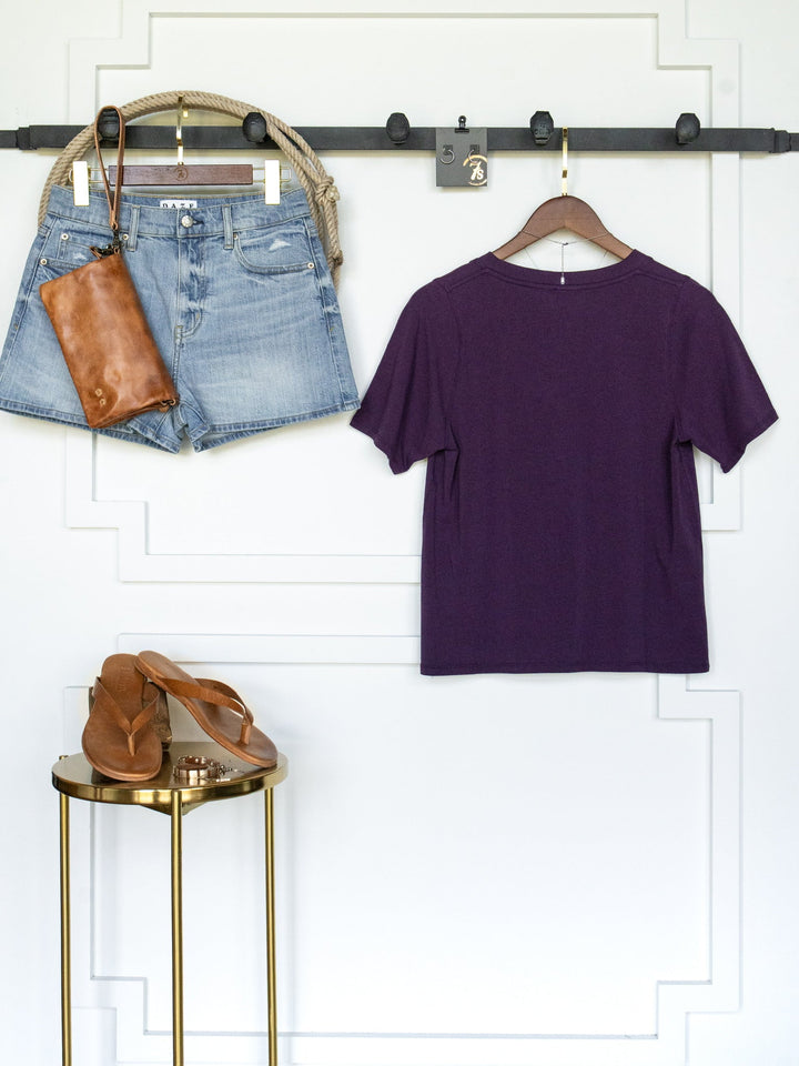 The Wymore Eggplant Basic Tee by Savannah Sevens western life{&}style hangs on a hanger, paired with denim shorts, a brown leather clutch, and sandals—all arranged neatly against a white wall with decorative trim.