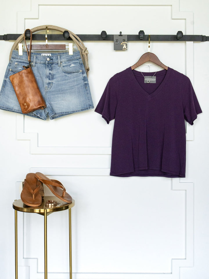 The Wymore Eggplant Basic Tee by Savannah Sevens western life{&}style, paired with denim shorts, a brown leather purse, and brown sandals, is neatly arranged against a white wall with panel detailing and a gold-colored stool.