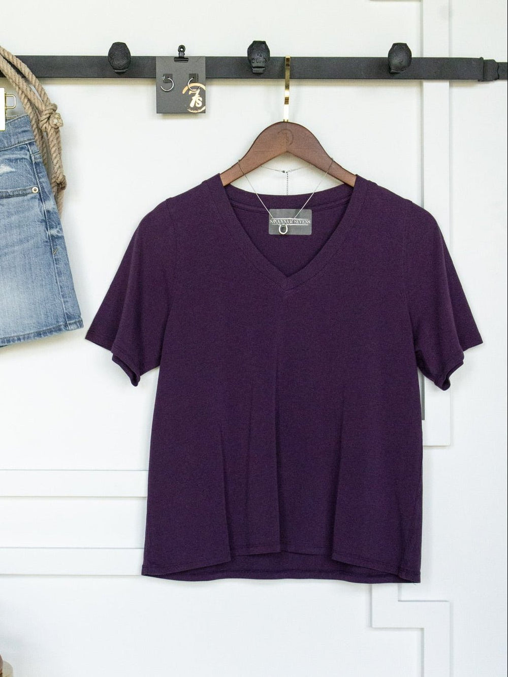 The Wymore Eggplant Basic Tee by Savannah Sevens western life{&}style, featuring a V-neck and short sleeves in pima cotton, hangs on a wooden hanger against a white wall, with part of blue denim shorts visible to the left.