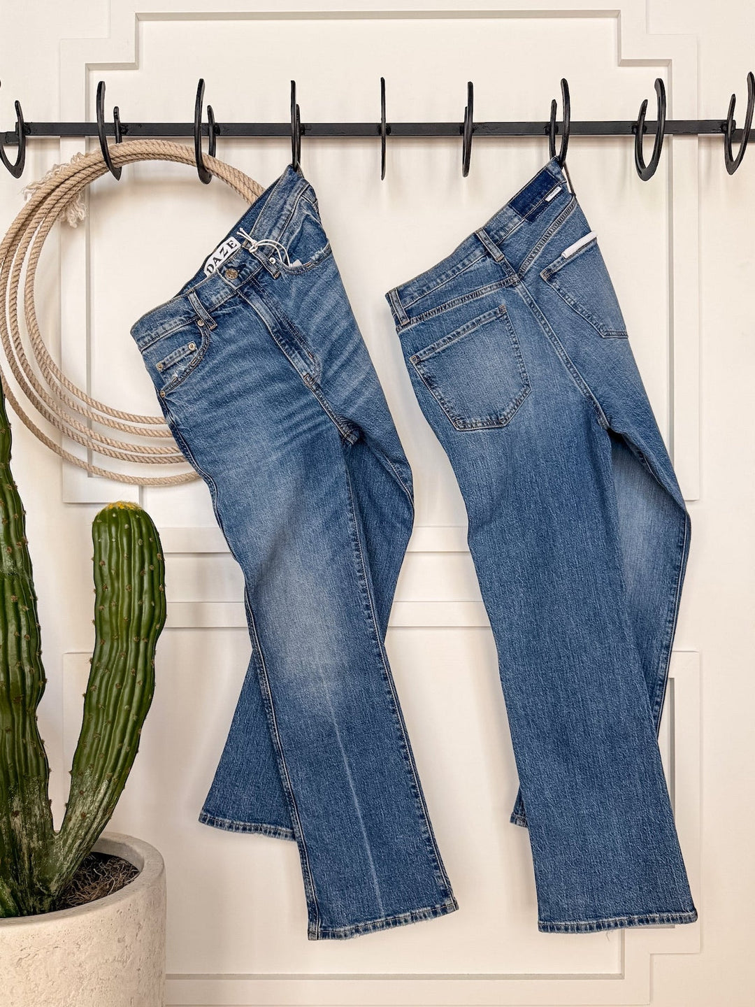 Two pairs of The Wynn High Rise Ankle Jean by Savannah Sevens western life{&}style hang from black hooks on a white paneled door, with a green cactus in a white pot and a coiled rope beside the jeans.