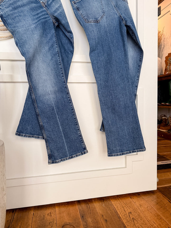 Two pairs of The Wynn High Rise Ankle Jean by Savannah Sevens western life{&}style in medium wash hang side by side on a white paneled wall, with a wooden floor and part of a cozy, warmly lit room in the background.