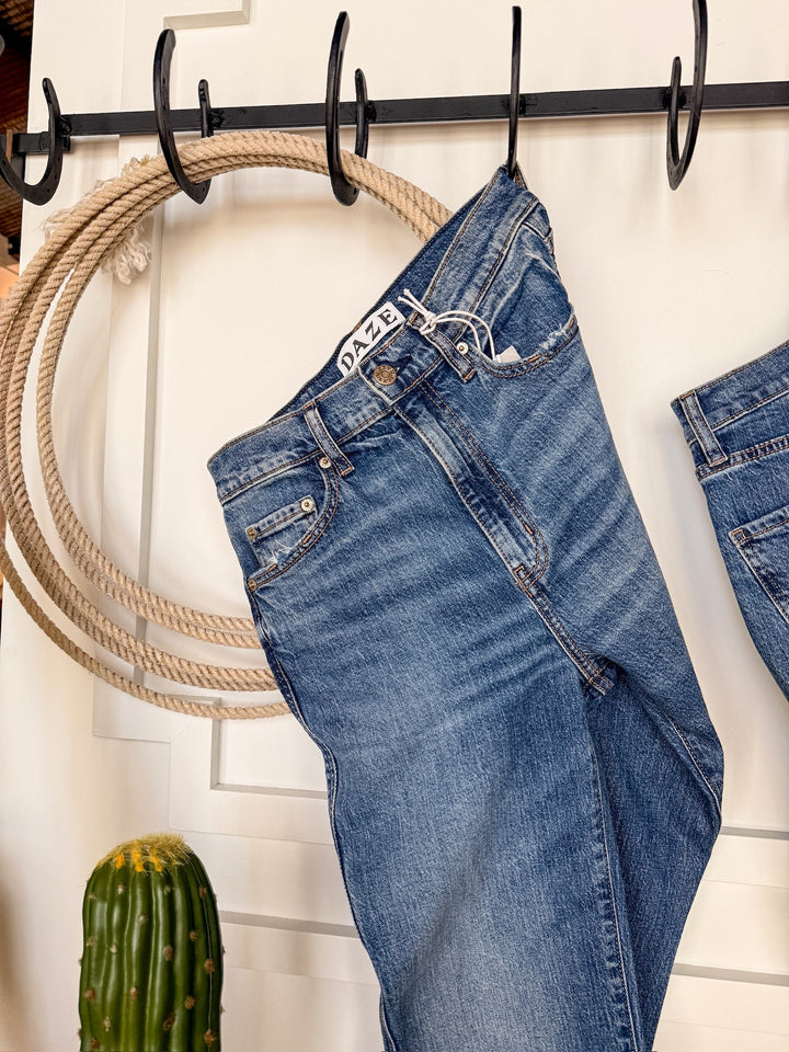 The Wynn High Rise Ankle Jean by Savannah Sevens western life{&}style in medium wash denim hangs on a black hook before a coiled rope, with a cactus peeking into the lower left corner.