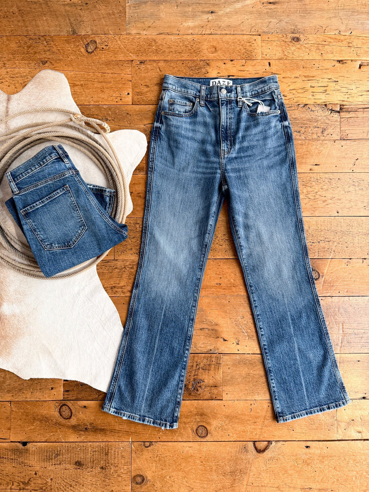 A pair of The Wynn High Rise Ankle Jeans by Savannah Sevens western life{&}style lies flat on a wooden floor beside folded Daze jeans, a coiled rope, and a cream-colored rug.