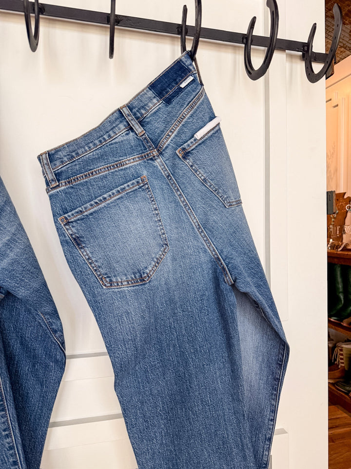 A pair of The Wynn High Rise Ankle Jean by Savannah Sevens western life{&}style in medium wash hangs on a black hook rack attached to a white door, displayed from the back to show its pockets and waistband.