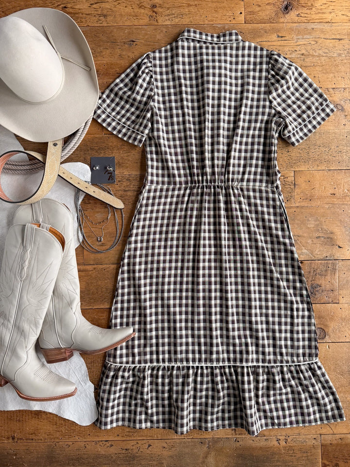 The Wynnewood Espresso Gingham Pearl Snap Dress from Savannah Sevens western life{&}style is laid out on a wooden floor, accompanied by a white cowboy hat, cream cowboy boots, a white belt, layered necklaces, and silver earrings.