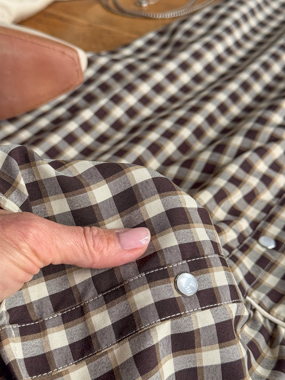 A hand with light pink nail polish holds a section of the Savannah Sevens western life{&}style Wynnewood Espresso Gingham Pearl Snap Dress, featuring classic brown and cream gingham plaid fabric with distinctive white pearl snap buttons that evoke a western yoke style. In the background, a tan bag and a silver chain are partially visible.