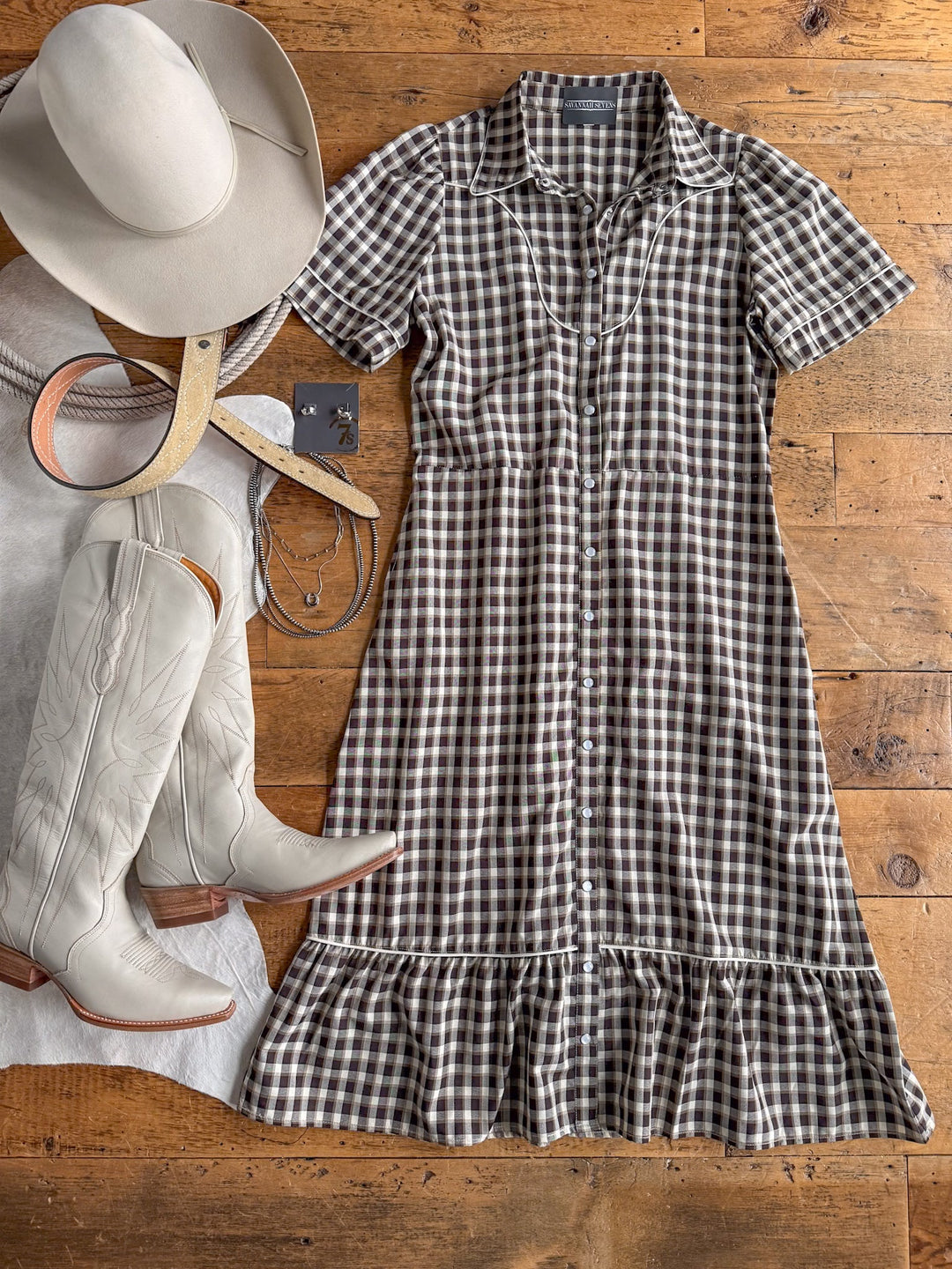 The Wynnewood Espresso Gingham Pearl Snap Dress by Savannah Sevens western life{&}style is styled with a white cowboy hat, tan belt, white cowboy boots, and a necklace on a wooden floor.