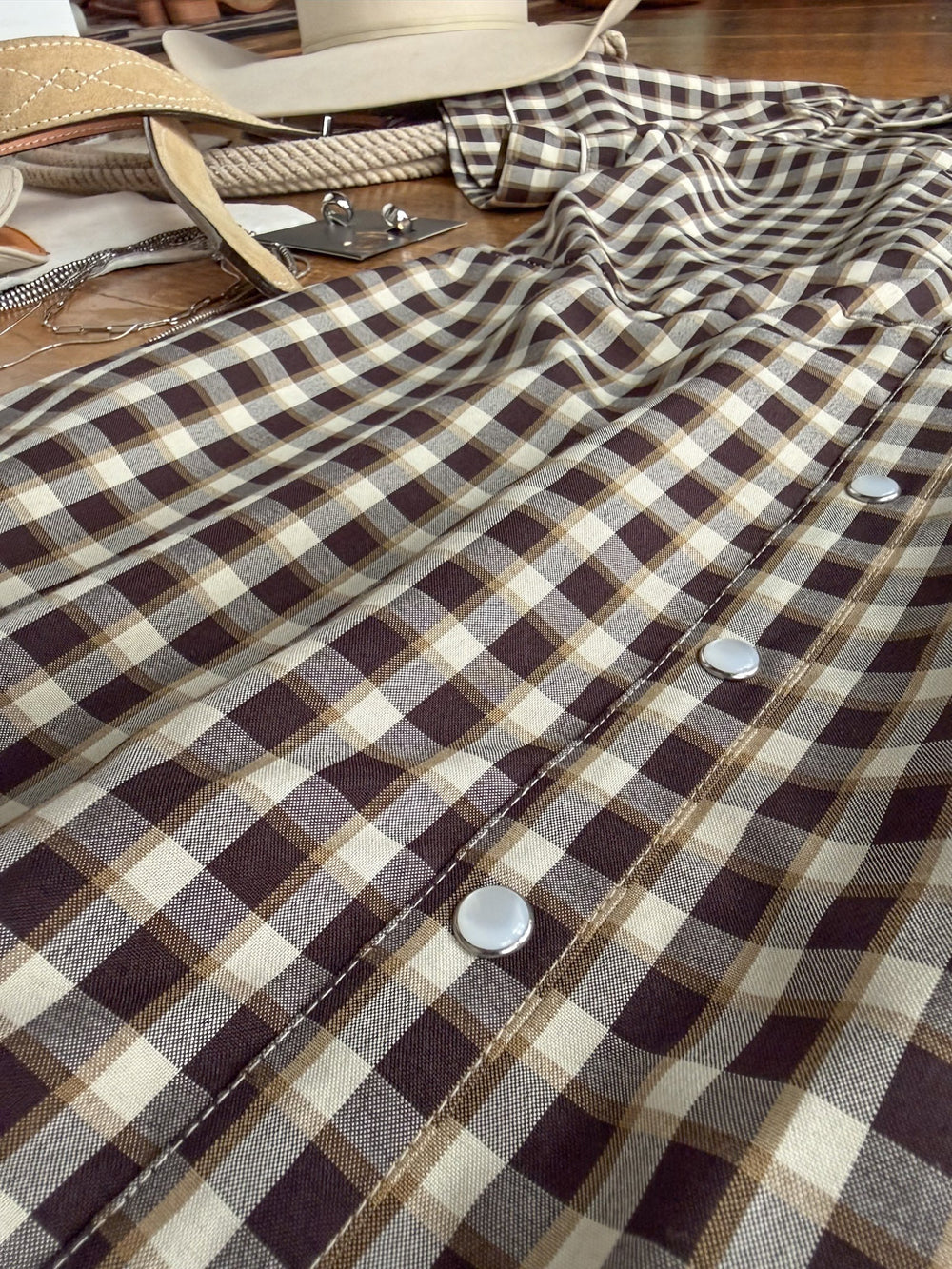 A close-up of the Wynnewood Espresso Gingham Pearl Snap Dress by Savannah Sevens western life{&}style showcases its brown and tan gingham pattern with pearl snap closures and a subtle western yoke detail, laid out on a surface. In the background, a tan belt, a hat, and metal accessories are partially visible.