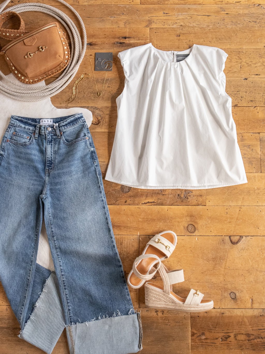 Flat lay featuring the Savannah Sevens western life{&}style Yampa White Sleeveless Top, light blue cuffed relaxed-fit jeans, tan wedge sandals, a tan crossbody bag, gold jewelry, and a coiled rope on a wooden floor.