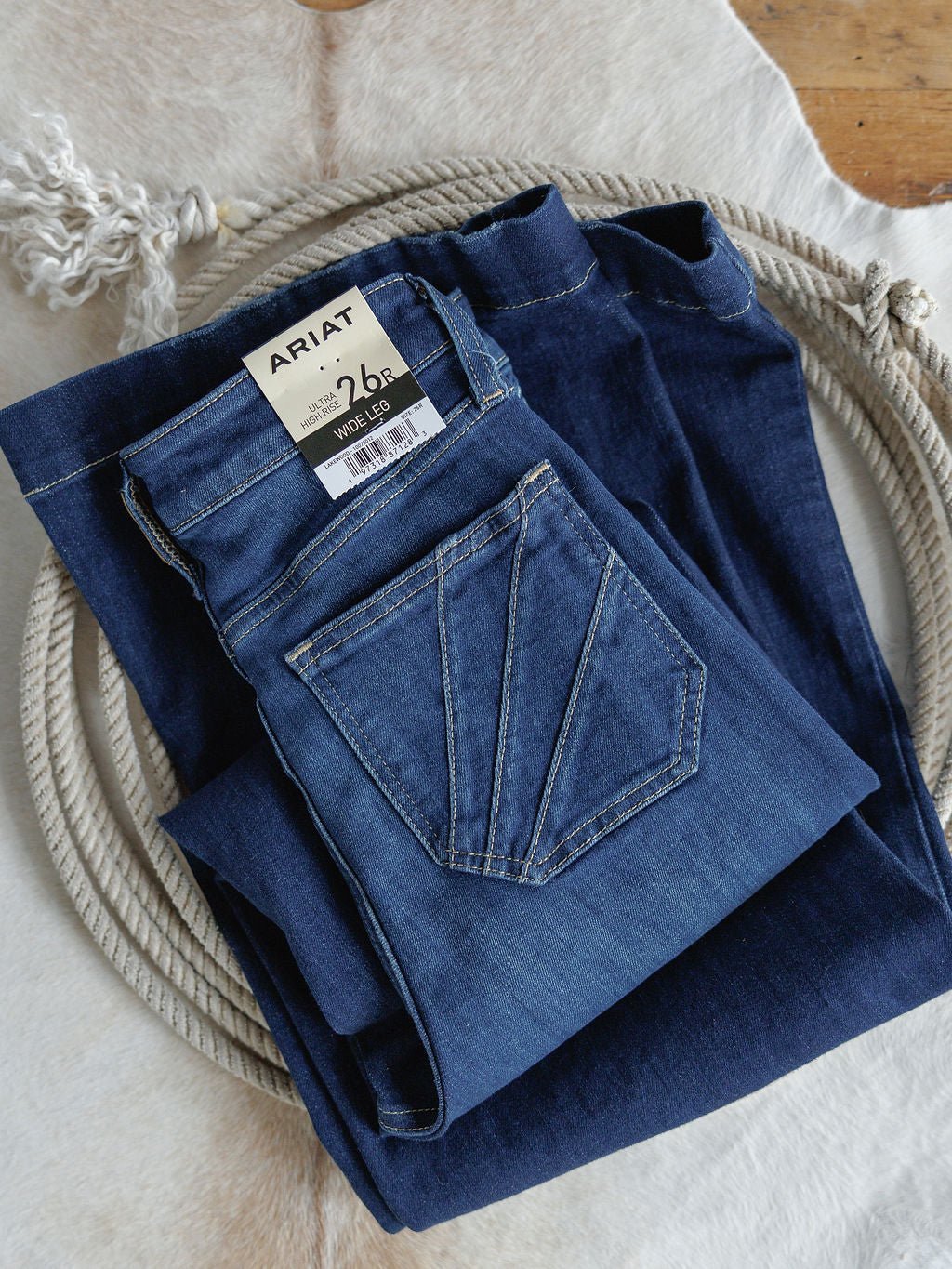 A folded pair of Ariat’s The Yates High Rise Wide Leg Jeans with a visible brand tag rests on a coiled rope atop a light cowhide rug. These jeans feature an ultra high rise, wide leg fit, and stylish medium dark wash.