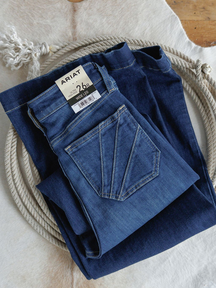 A folded pair of Ariat’s The Yates High Rise Wide Leg Jeans with a visible brand tag rests on a coiled rope atop a light cowhide rug. These jeans feature an ultra high rise, wide leg fit, and stylish medium dark wash.