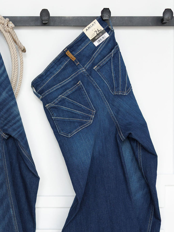 A pair of Ariat “The Yates High Rise Wide Leg Jean” in a medium dark wash hangs on a black hook rack with a size 26R tag. Another denim piece and a rope are partially visible beside the jeans.