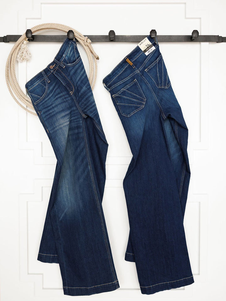 Two pairs of Ariat The Yates High Rise Wide Leg Jeans in a medium dark wash hang side by side on a black wall rack with looped rope, showing subtle fading and contrast stitching against a white, decorative-paneled background.