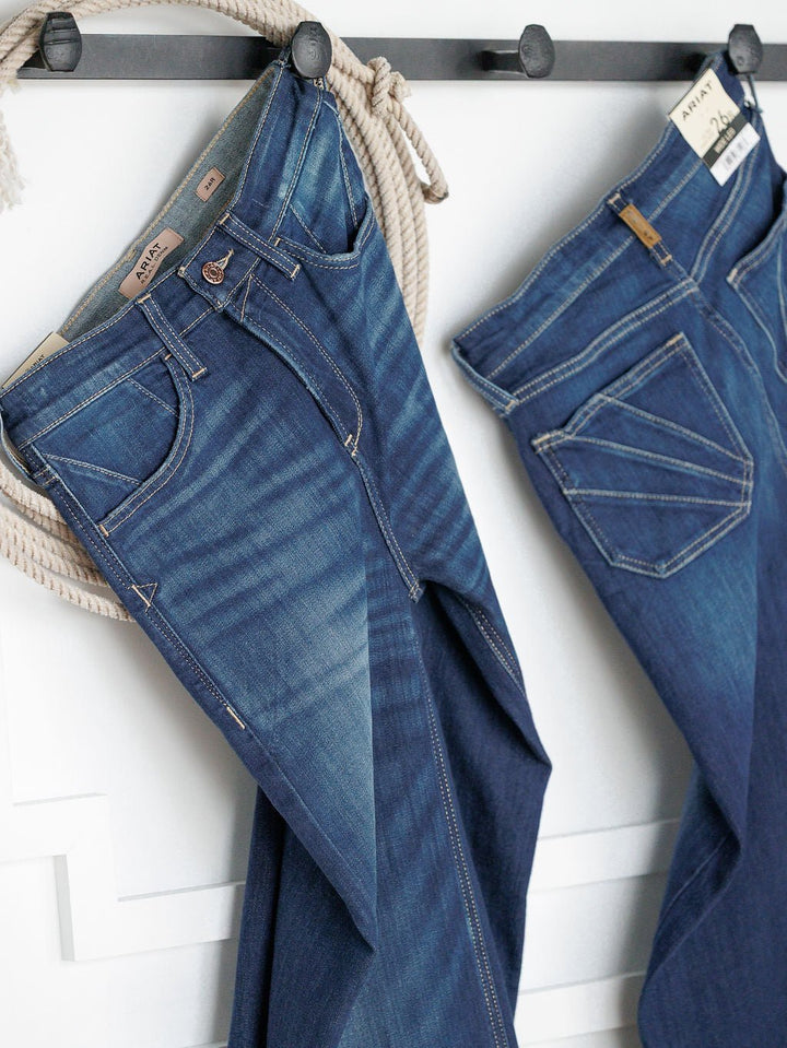 Two pairs of Ariat The Yates High Rise Wide Leg Jeans in medium dark wash hang on black hooks against a white wall, with a coiled rope draped over the top hook. Both showcase white stitching and brown leather Ariat brand patches.