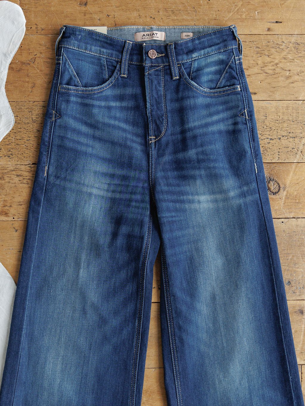 The Yates High Rise Wide Leg Jean by Ariat, in a medium dark wash with a faded, worn-in look, features ultra high rise style, front pockets, belt loops, and visible stitching. Jeans are shown laid flat on a wooden floor.