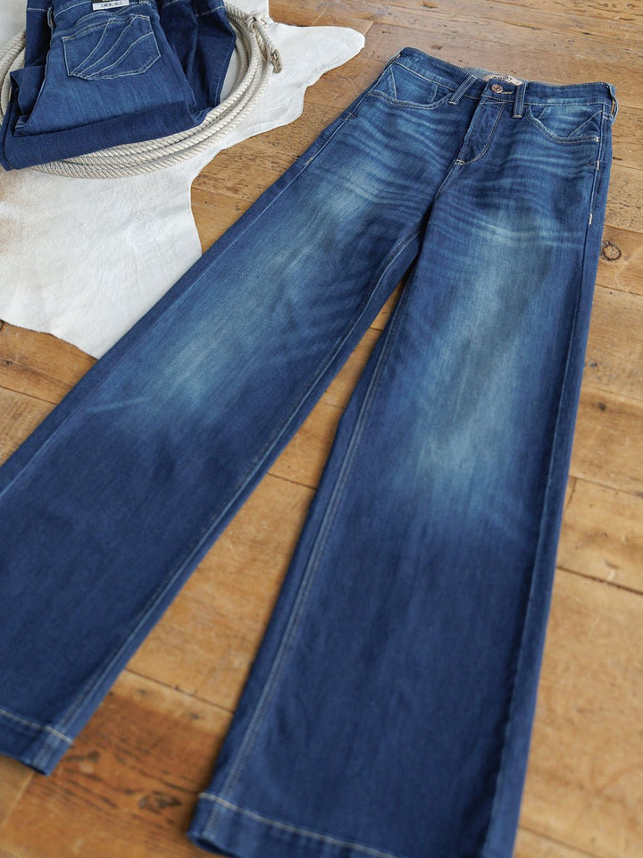 The Yates High Rise Wide Leg Jean by Ariat in a medium dark wash is laid flat on a wooden floor, with another pair of folded Ariat denim and a white cloth in the background. Faded details accent the thighs and seams.