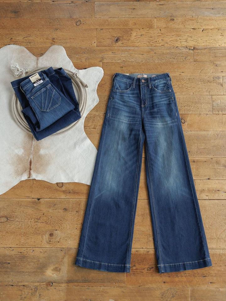 The Yates High Rise Wide Leg Jean by Ariat, in a medium dark wash, is laid flat on a wooden floor beside a folded stack of jeans on a white fur rug, showcasing the denim texture and faded details.