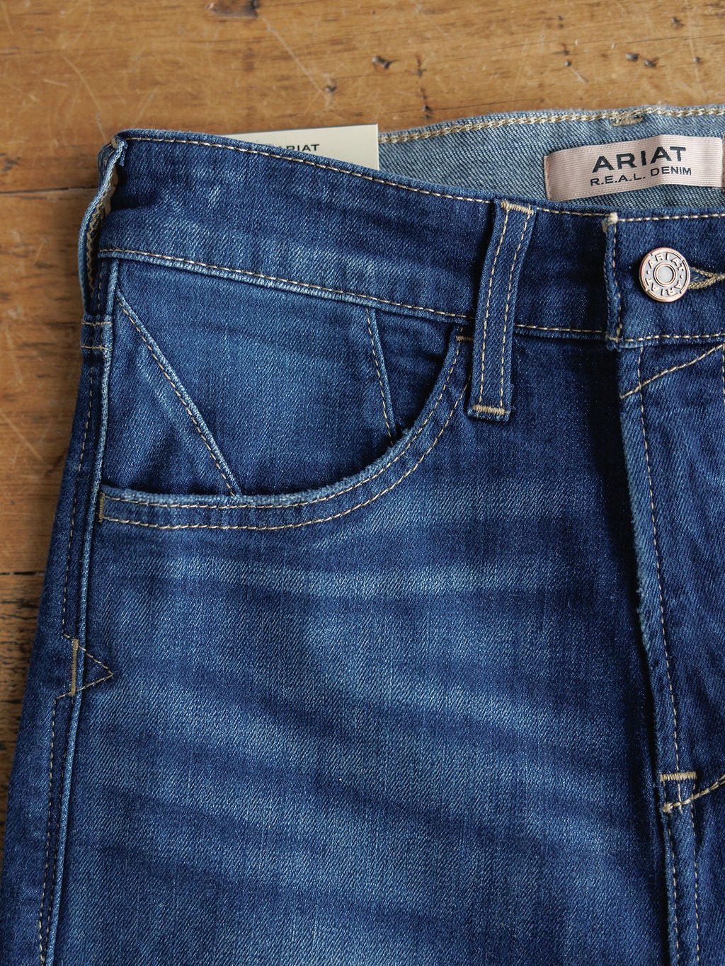 Close-up of the top of Ariat's The Yates High Rise Wide Leg Jean in medium dark blue denim on wood, showing the front pocket, belt loops, and a brown leather Ariat label inside the waistband.