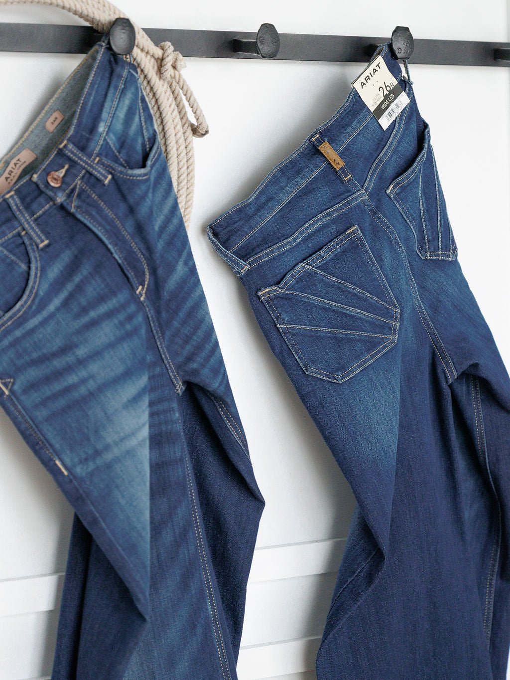Two pairs of The Yates High Rise Wide Leg Jeans by Ariat hang on black hooks against a white wall, both displaying medium dark wash fading, visible stitching, and “ARIAT” labels.