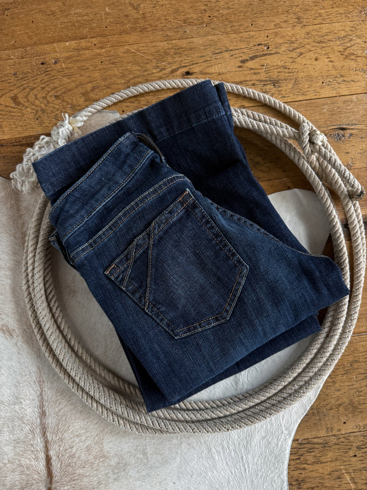 A pair of folded Savannah Sevens western life{&}style Yoakum Perfect Rise Trouser Jeans *Exclusive* in a medium dark wash rests on a coiled rope atop a white cowhide, placed on a wooden floor.
