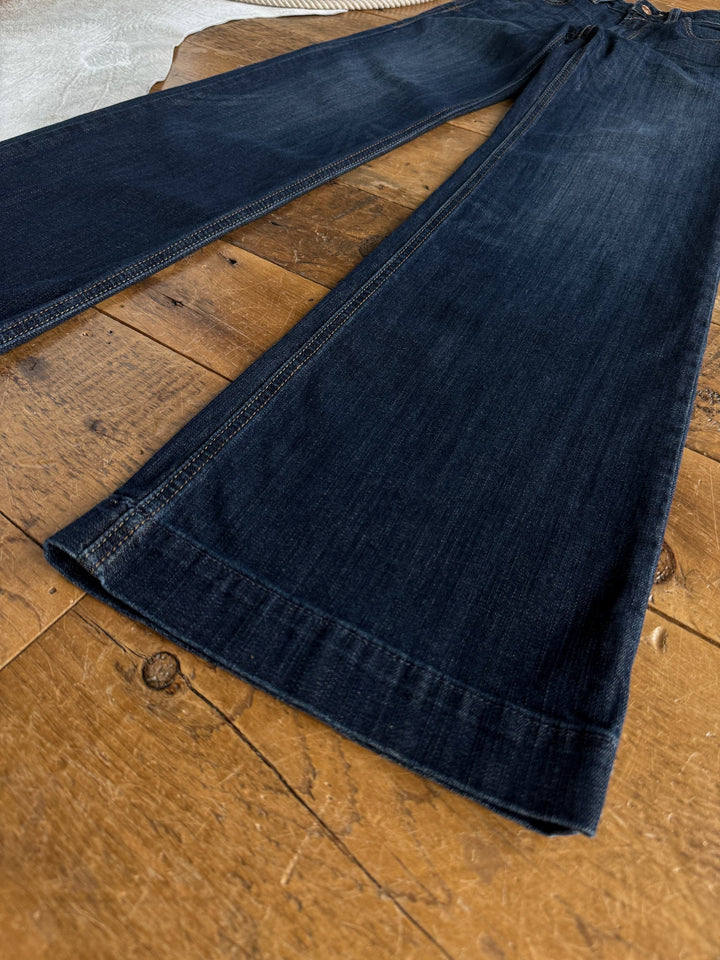 Savannah Sevens western life{&}style’s Yoakum Perfect Rise Trouser Jean *Exclusive* in a medium dark wash is laid flat on a rustic wooden surface. The image highlights the wide leg hems and perfect rise, focusing on the lower half of the jeans.