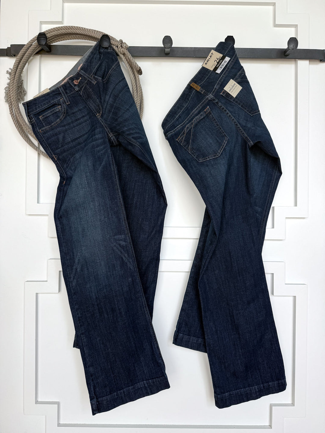 Two pairs of “Yoakum Perfect Rise Trouser Jean *Exclusive*” by Savannah Sevens western life{&}style, in a medium dark wash, hang on a black bar against a white panel wall. One pair still has tags attached, and a gray rope is looped over the bar on the left side.