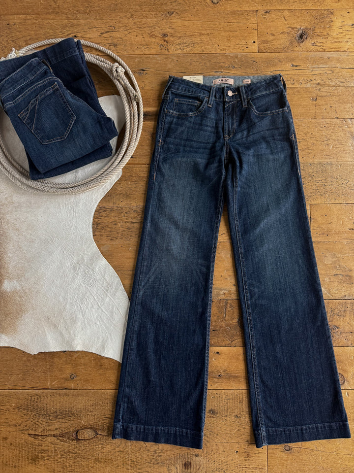 A pair of Savannah Sevens western life{&}style Yoakum Perfect Rise Trouser Jeans in a medium dark wash lies flat on a wooden floor next to a folded pair of Ariat denim trousers, a coiled rope, and a light-colored animal hide.