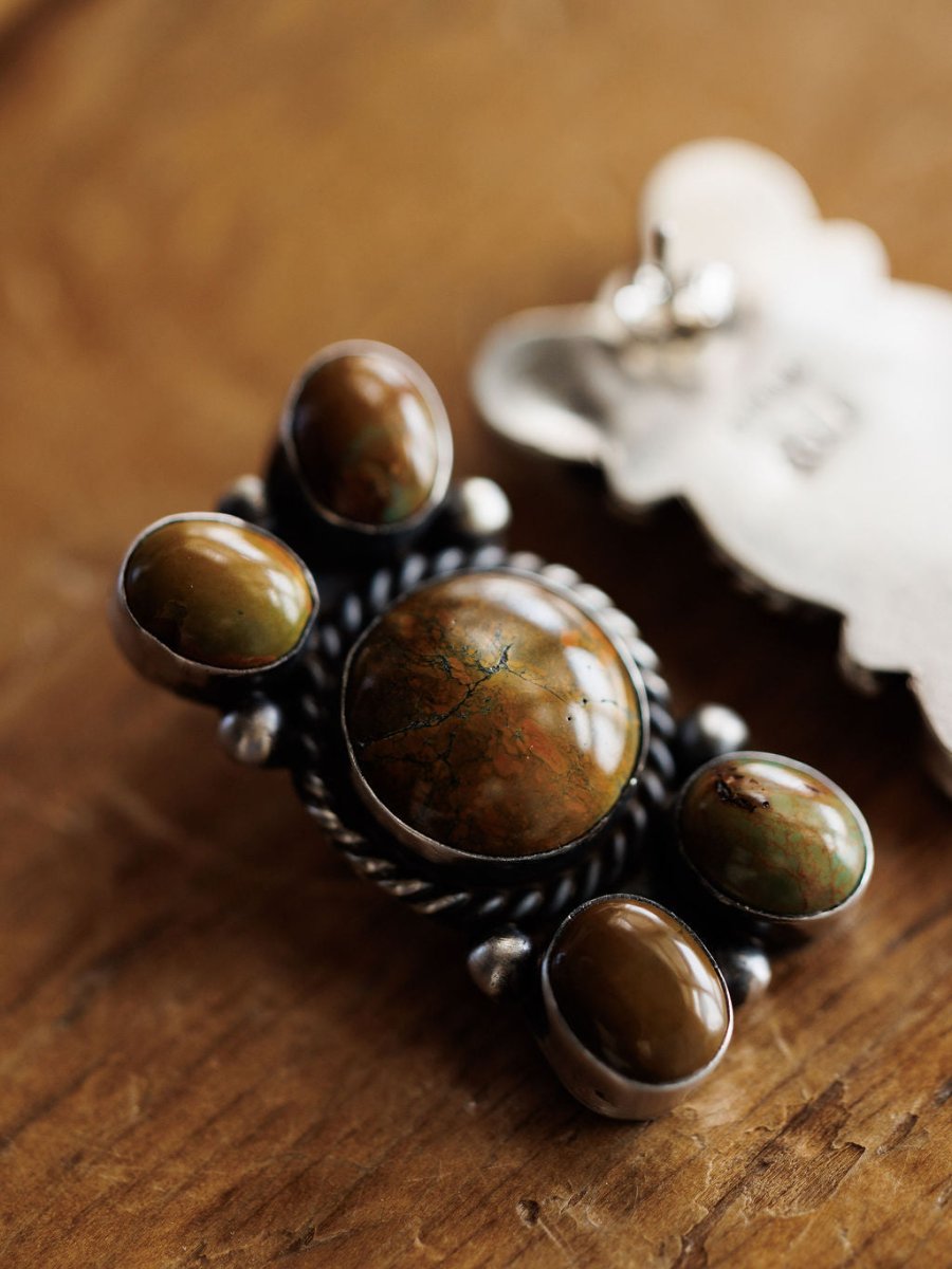 The Yuma Earrings by Savannah Sevens western life{&}style feature handcrafted sterling silver with five polished brown and green stones in ornate metalwork, beautifully displayed on a wooden surface.