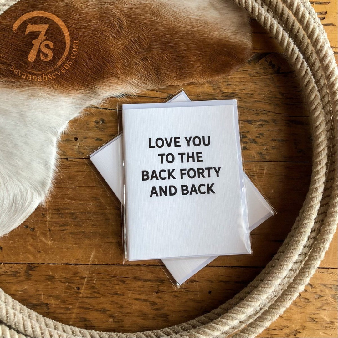 The Back Forty Card by Hungry Canyon, featuring "LOVE YOU TO THE BACK FORTY AND BACK" on linen paper, sits on wood with a cowhide patch and rope nearby. The card is blank inside. The savannahseven.com logo appears in the top left corner.