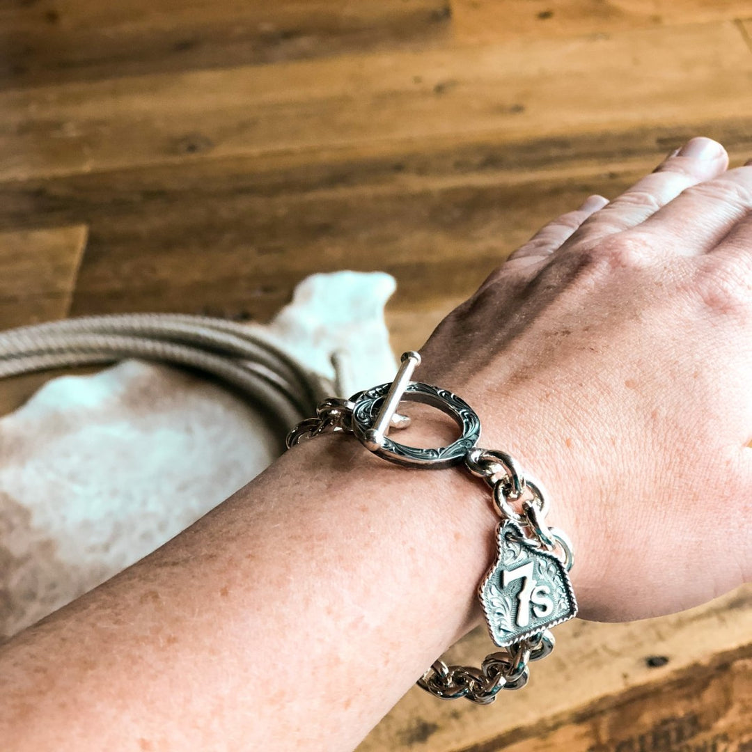 A hand wears a Savannah Sevens western life{&}style Boca Raton Bracelet, featuring sterling silver with a toggle clasp and engraved 7s charm. The background includes wood, coiled rope, and cowhide.
