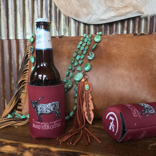 A Brand Your Cattle Koozie {cranberry} from Savannah Sevens western life{&}style holds a beer bottle on a wooden surface, with a turquoise bead necklace, leather pouch, and another koozie adding to the Western Chic style.